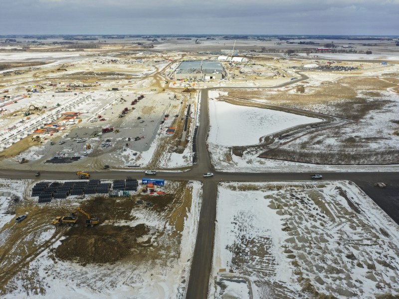 An aerial view shows a large construction site with cranes, heavy equipment and materials surrounded by snow-covered fields and intersecting roads.