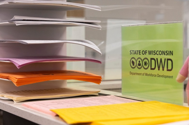Stacks of paper of varying colors sit on a counter and in rows in a document holder beside a light green sign with black letters reading “STATE OF WISCONSIN DWD Department of Workforce Development”