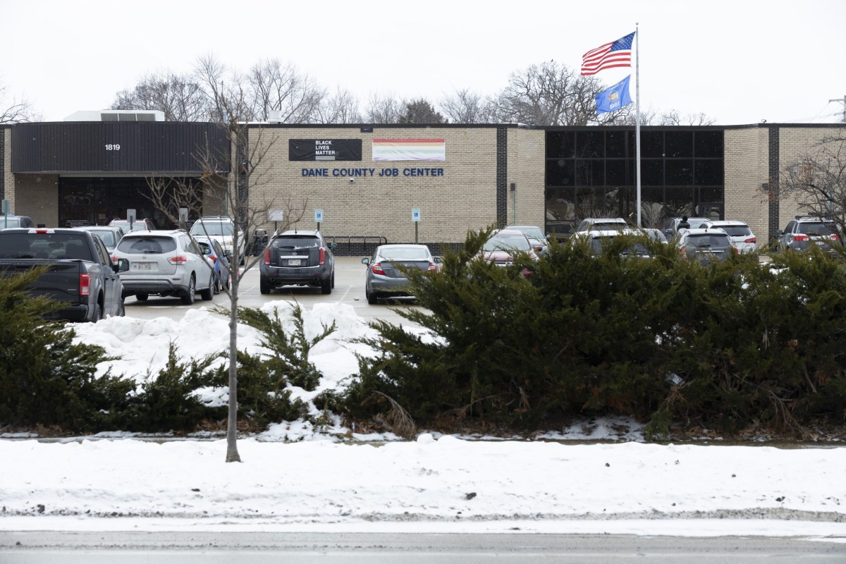 A brick building labeled “DANE COUNTY JOB CENTER” sits behind a parking lot with parked cars, shrubs in front, snow on the ground, and U.S. and state flags flying on a pole.