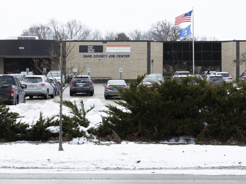 A brick building labeled “DANE COUNTY JOB CENTER” sits behind a parking lot with parked cars, shrubs in front, snow on the ground, and U.S. and state flags flying on a pole.