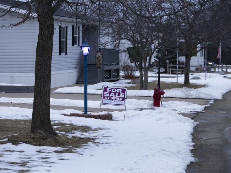 A "FOR SALE BY OWNER" sign stands in snow beside a sidewalk near a home, with a lit blue lamp post, leafless trees and a red fire hydrant.