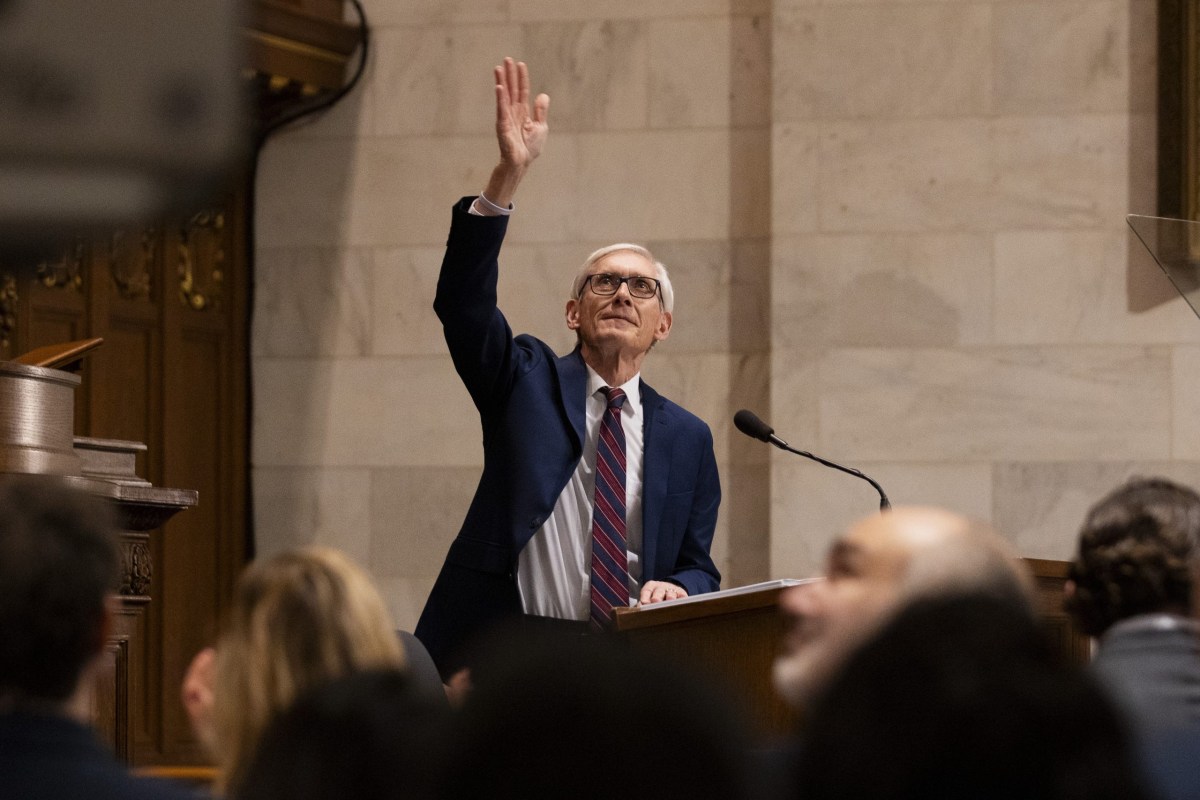 A person stands and raises a hand at a podium with a microphone in a marble-walled room, with other people sitting in the foreground.