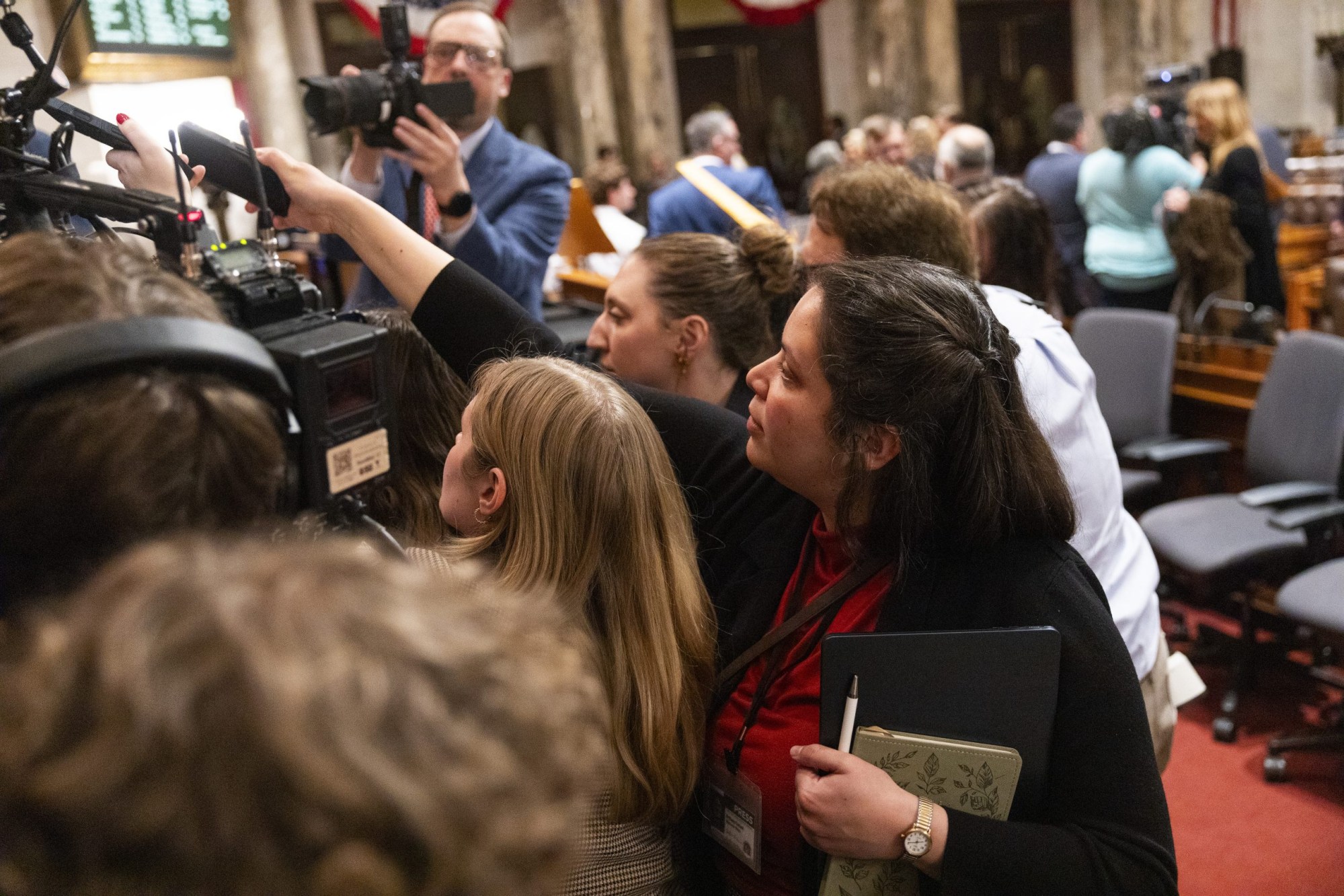 People gather closely around cameras and microphones in a room while a person holds a notebook, pen and smartphone in the foreground.