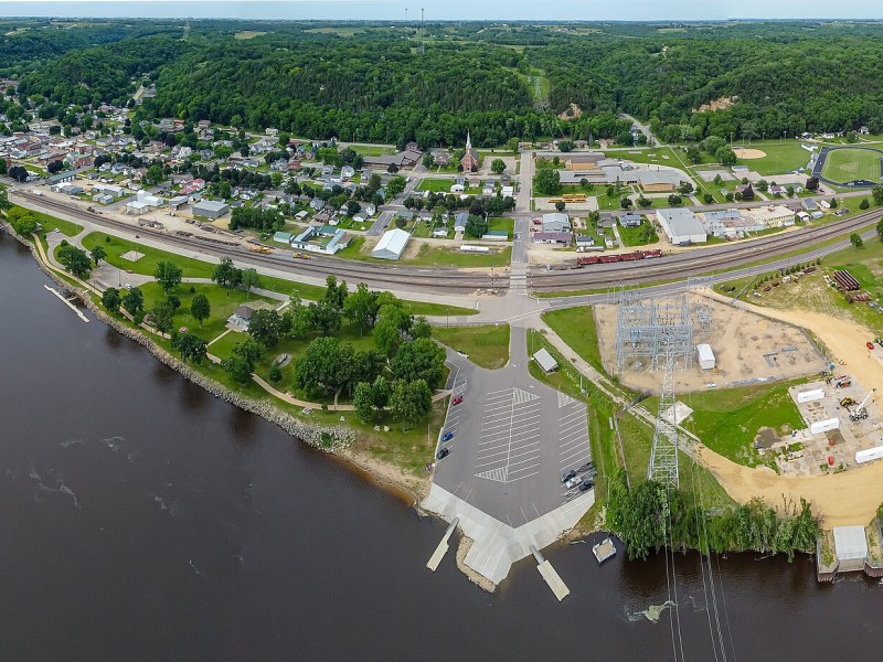 An aerial view shows a town along a wide river, with homes, roads, railroad tracks, green hills, and a construction site with utility structures near the shoreline.