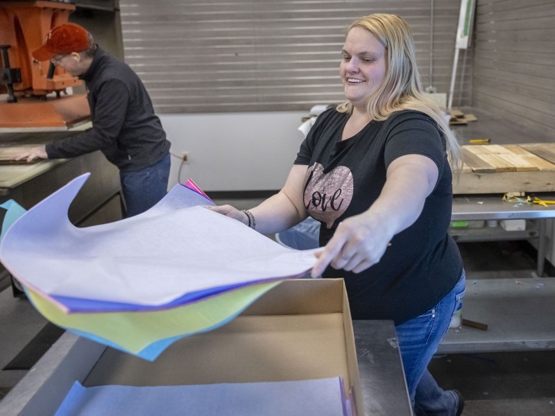 A person places sheets of colorful material into a cardboard box at a worktable while another person stands at another worktable in the background.