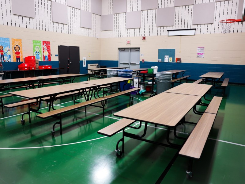 An empty room with long tables, attached benches and a green floor, with colorful posters and a basketball hoop on the walls.