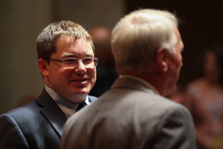 Two people in suits stand near each other, with one person smiling and wearing glasses and looking at the other, who is seen from behind.