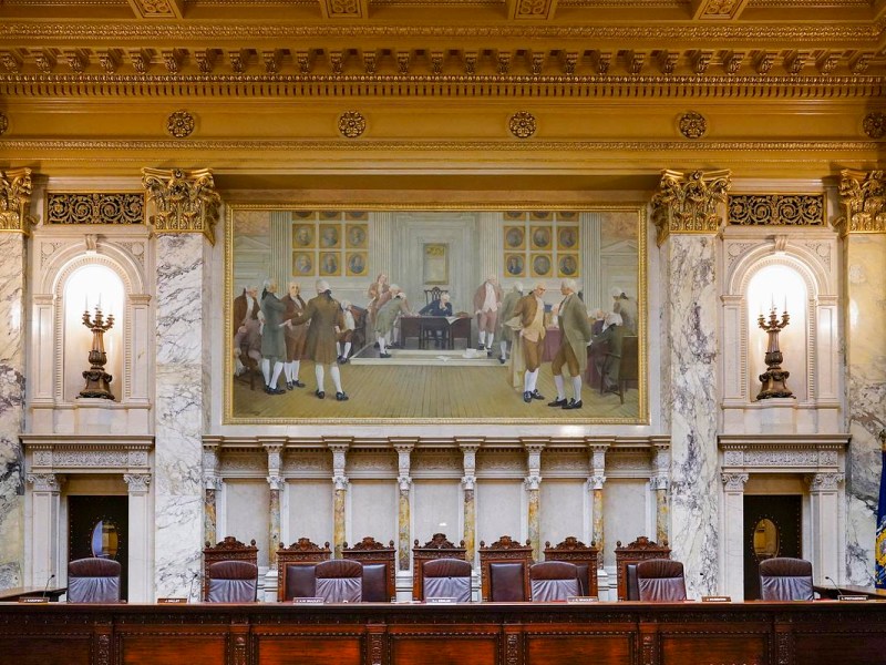A row of wooden chairs and microphones sits beneath marble walls and a large framed painting of people gathered in a historical interior.