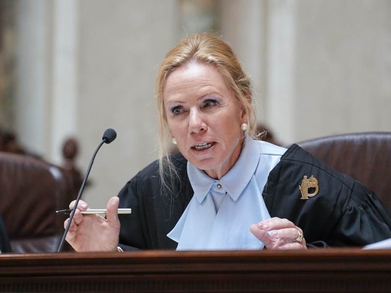 A person in a black and blue robe sits in a chair and holds a pen near a microphone.