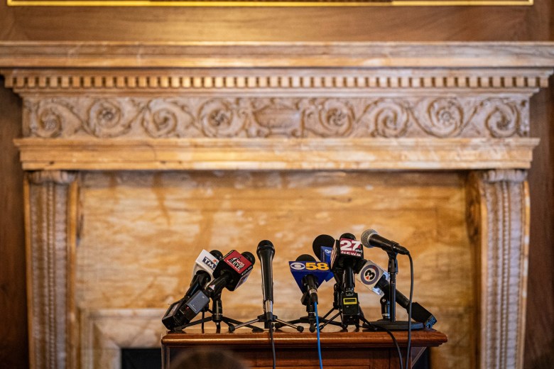 A cluster of microphones with news station logos sits on a wooden podium in front of an ornate mantel.