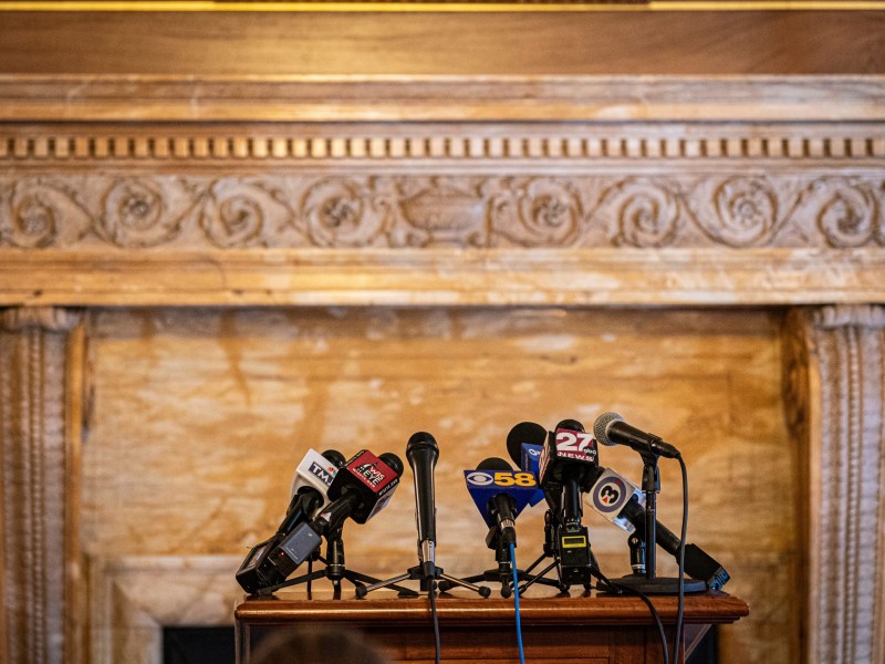A cluster of microphones with news station logos sits on a wooden podium in front of an ornate mantel.
