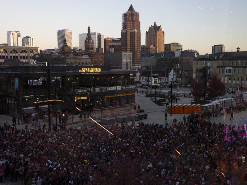 A large crowd gathers in a downtown plaza near a building with a sign reading "THE NEW FASHIONED," with high-rise buildings and a city skyline in the background.