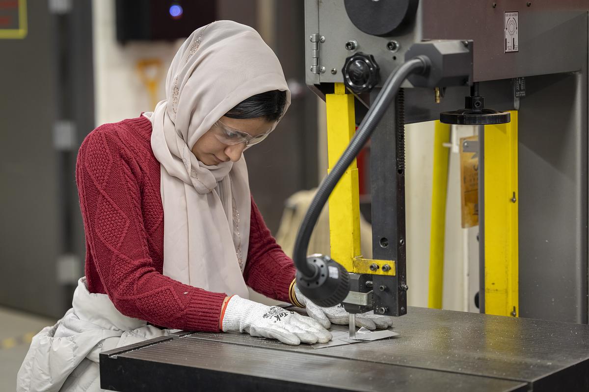 A person wearing gloves, a red sweater, a head covering and safety glasses positions a metal piece under a vertical machine on a worktable in an industrial room.
