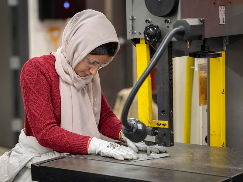 A person wearing gloves, a red sweater, a head covering and safety glasses positions a metal piece under a vertical machine on a worktable in an industrial room.