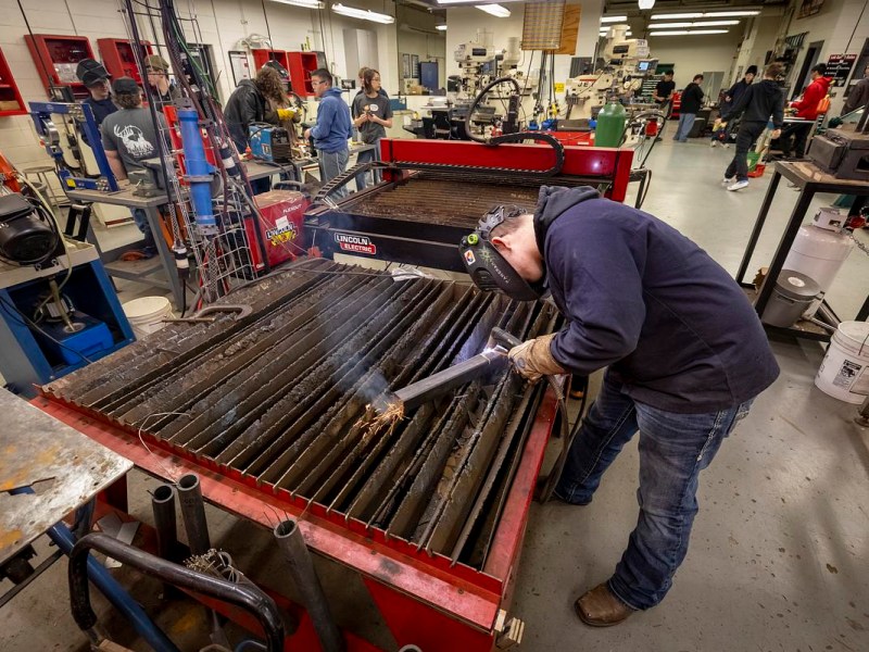 A person wearing gloves and a welding helmet uses a torch on a metal sheet atop a large table, with sparks flying and several other people and machines visible in a workshop.
