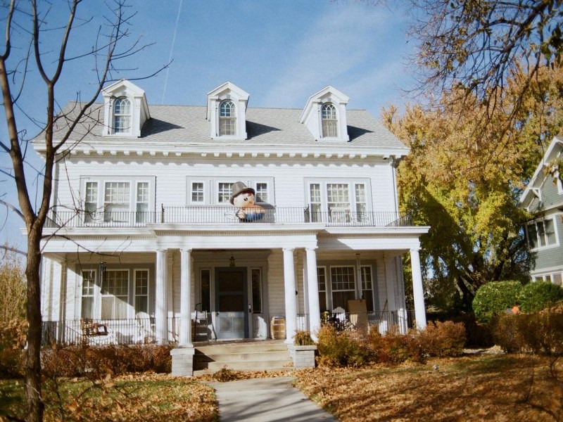 A large white house with columns and dormer windows has an inflatable figure wearing a hat on an upper balcony, with autumn leaves covering the lawn.