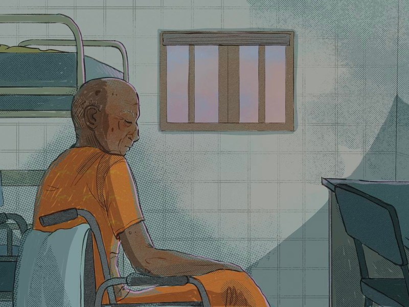 Person wearing orange clothes sits in a wheelchair in a prison cell.