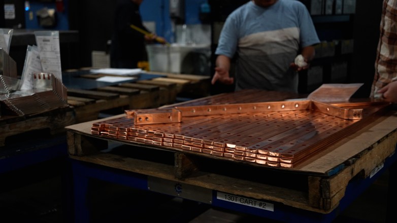 Stacks of copper bars with drilled holes sit on a wooden pallet in a workshop, with a person standing nearby in the background.