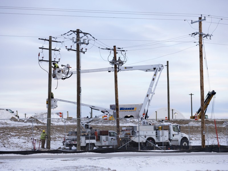 People in raised bucket trucks work on utility poles and overhead power lines behind a chain-link fence, with snow on the ground and equipment vehicles parked nearby.