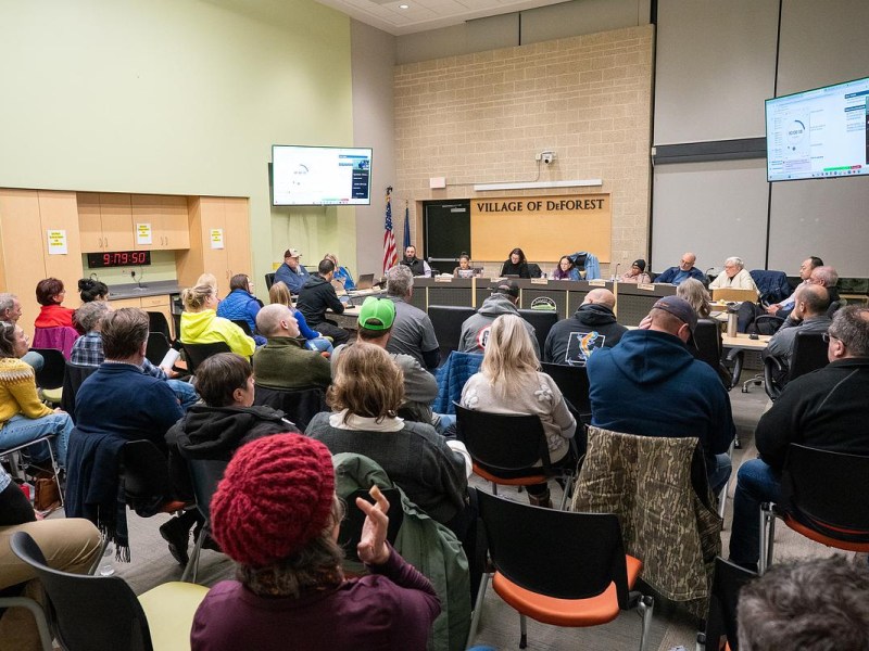 People sit in chairs facing a long desk in a room, with people seated behind microphones and a wall sign reading “Village of DeForest” above them.
