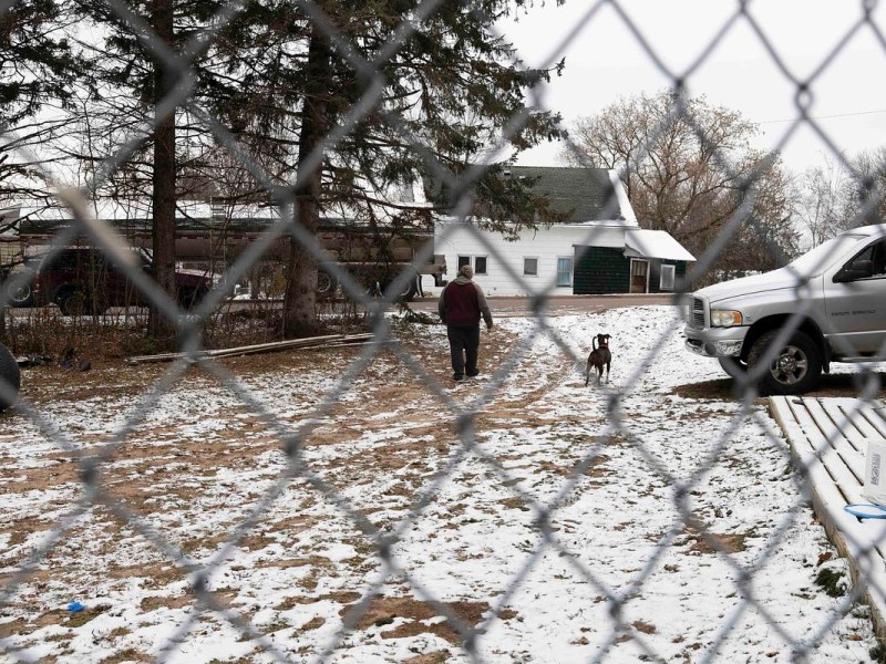 Man and dog walk on snow-covered ground away from fence.