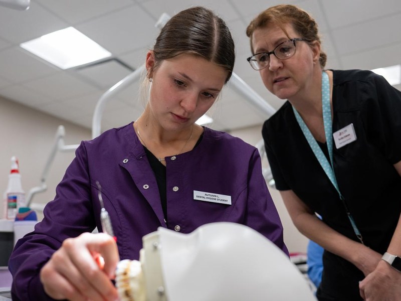 A person wearing a purple coat labeled "Dental Hygiene Student" works on a dental model while another person watches.
