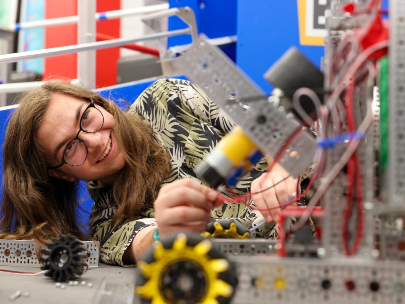 A person with glasses smiles while holding wires on a metal robot structure with wheels and gears on a worktable.