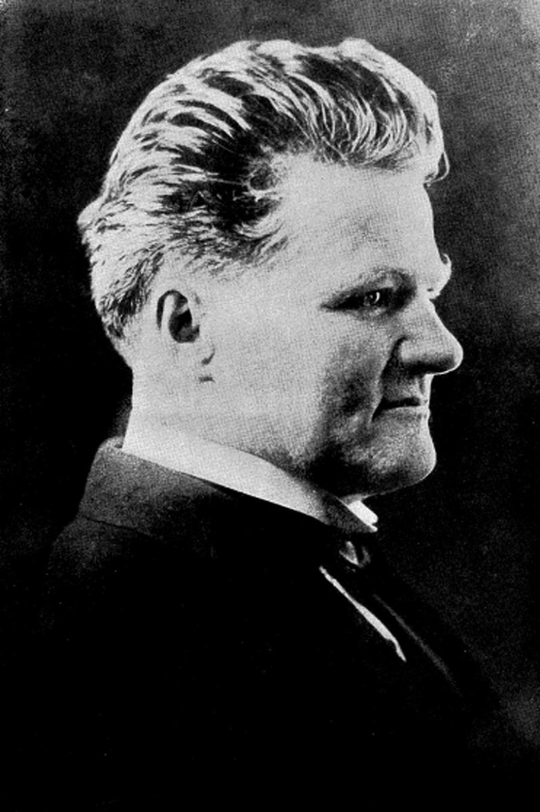 A person in a formal jacket is shown in a black-and-white side profile with short swept-back hair against a dark background.