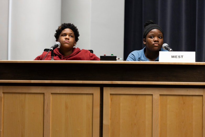 Two students sit next to each other behind a podium. A sign says "West" and a buzzer is shown.