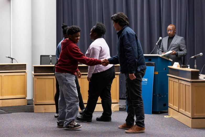 Two people shake hands in front of a dais with microphones while others stand nearby and a person at a podium holds papers against a backdrop of dark curtains