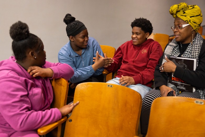 Four people sit in wooden auditorium seats talking; one gestures while another reaches toward their hand, and a person in a yellow headwrap holds a book reading "HISTORY"