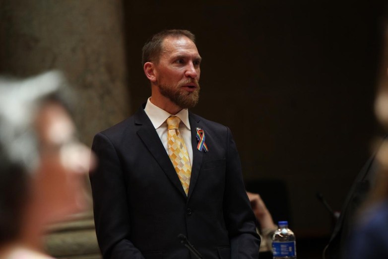 A person in a suit with a patterned tie and a multicolored ribbon on the lapel stands with a water bottle nearby.