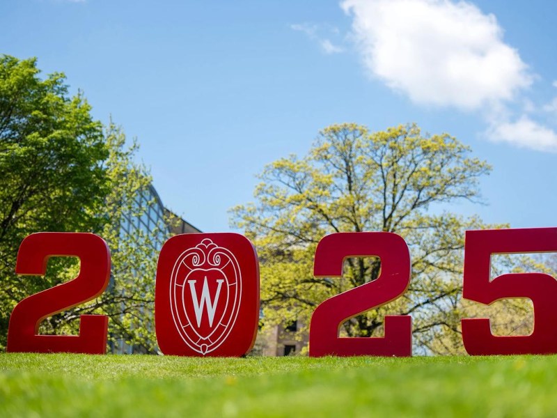 Large red numbers reading "2025" with a "W" emblem stand on a grassy area with trees and a building in the background under a blue sky.