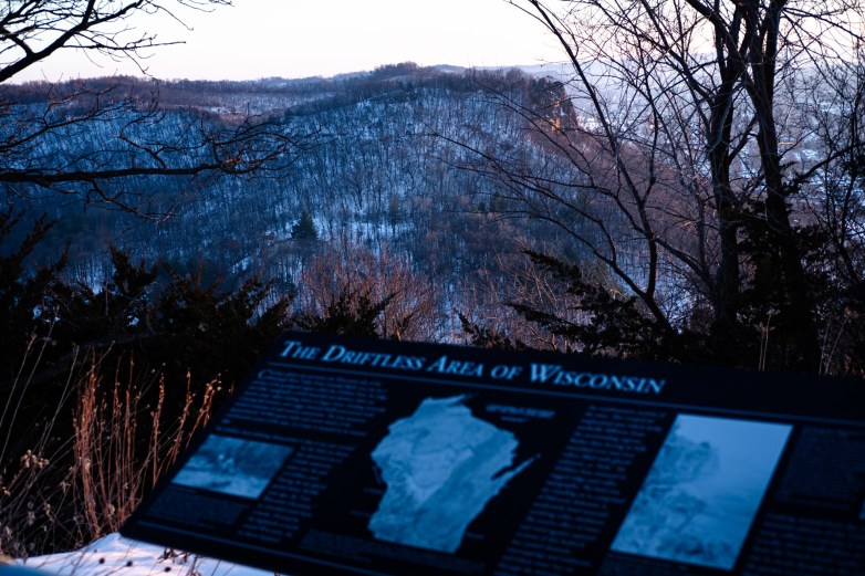An overlook sign reading "The Driftless Area of Wisconsin" stands in front of snow-covered wooded hills and a valley.