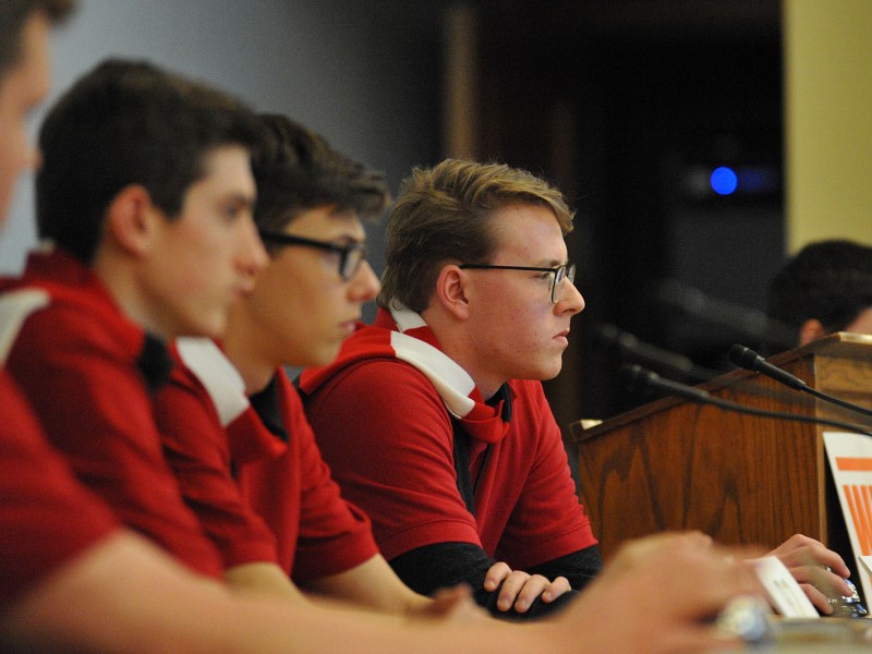 People wearing matching red shirts sit behind microphones at a long desk, facing forward in a panel setting.
