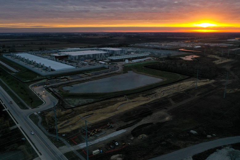 An aerial view of a large industrial complex next to a pond and surrounding construction areas at sunset, with orange light along the horizon under a cloudy sky.