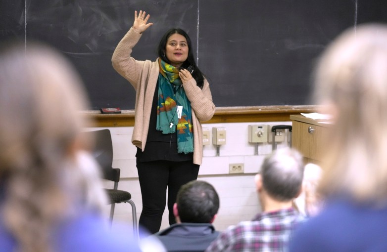 A person stands in front of a chalkboard holding a microphone and raising one hand, while other people sit facing the person.