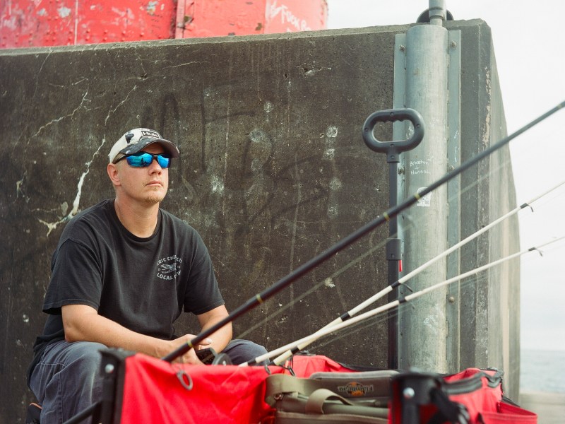 Man wearing sunglasses sits behind fishing poles.
