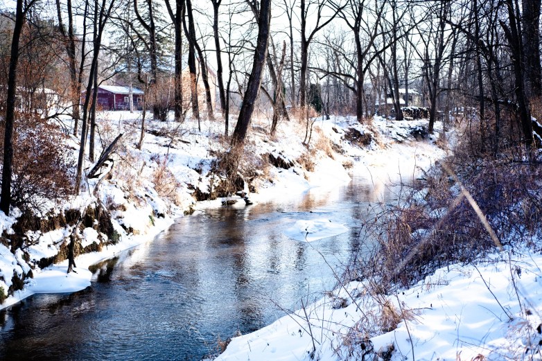 A creek flows between snow-covered banks lined with leafless trees and brush, with patches of ice along the water’s edge and houses and other buildings in the background.