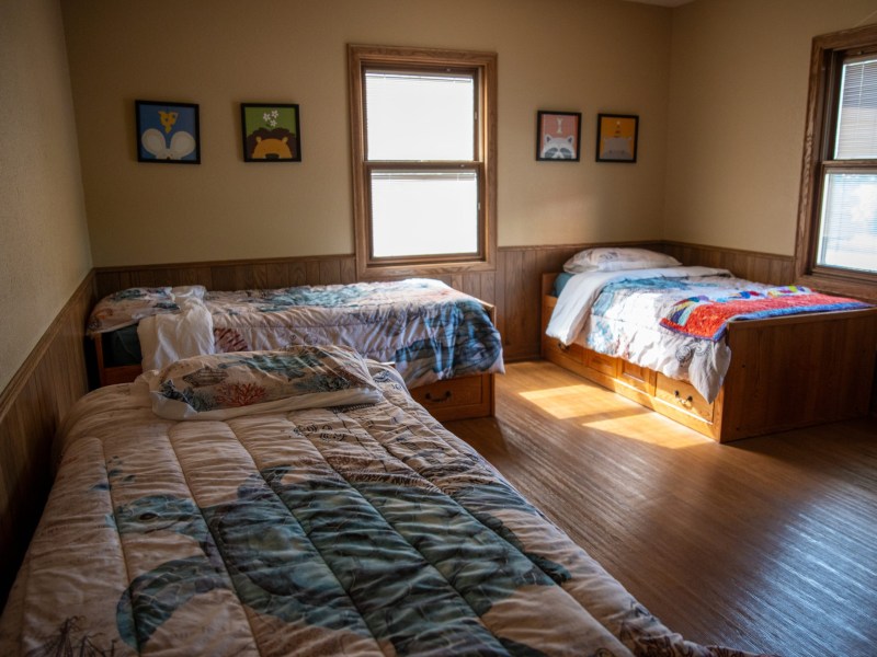 Three single beds with patterned quilts are next to the walls of a bedroom with wood paneling, hardwood floors, two windows, and small framed animal illustrations on a wall above the beds.
