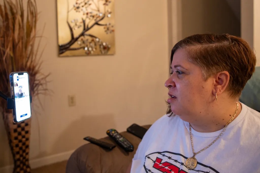 A person sits on a couch looking at a smartphone mounted on a stand displaying a video call screen, with remote controls on the arm of the couch and wall art in the background.
