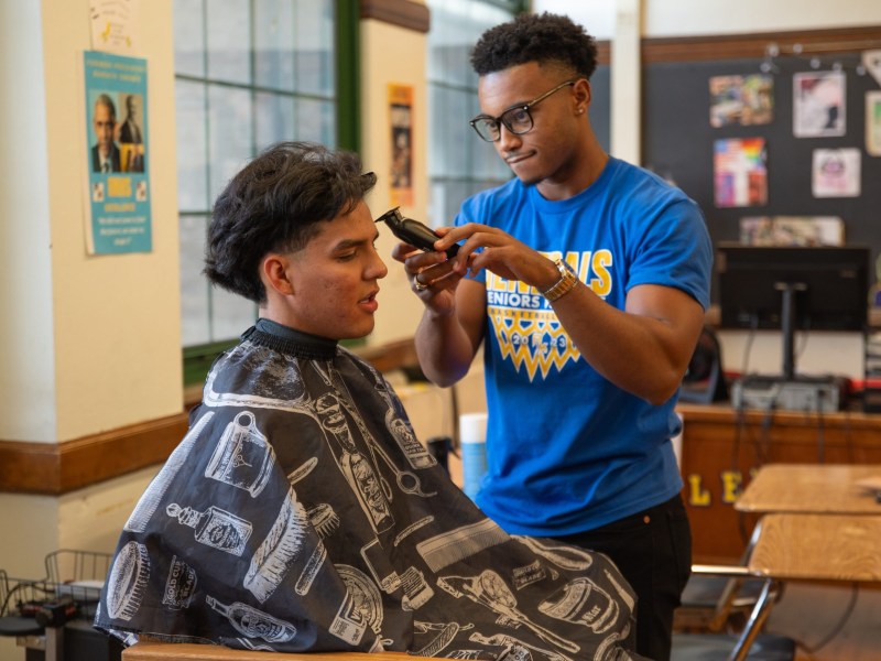 A person trims another person's hair with clippers in a room with desks, posters and a computer in the background.