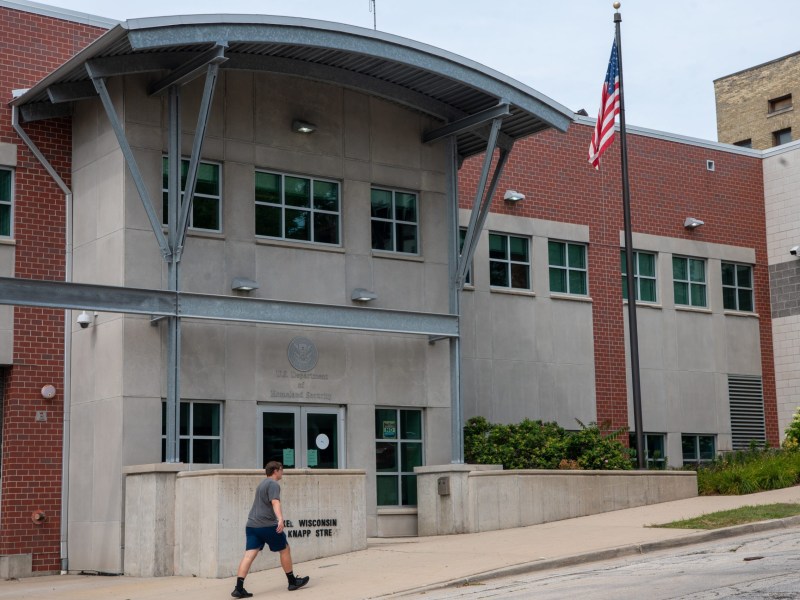 A person walks past a building with "U.S. Department of Homeland Security" above the entrance as an American flag flies on a pole in front of the building.