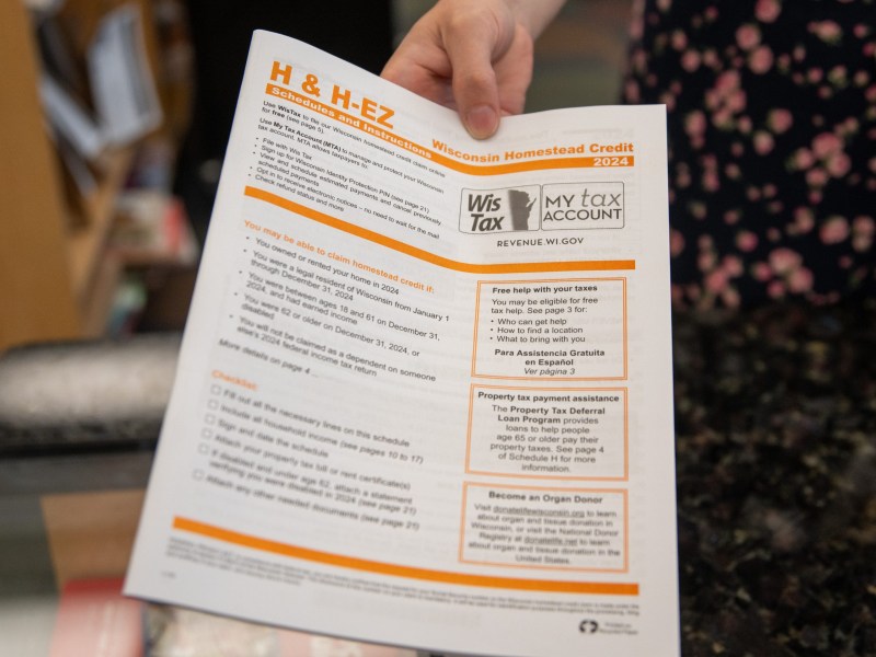 A person holds a Wisconsin Homestead Credit 2024 instruction form labeled "H & H-EZ" with "Wis Tax" and "MY tax ACCOUNT" logos visible near the top.