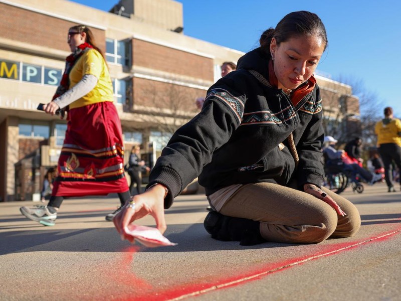 A person kneels on pavement spreading red sand as others walk nearby in front of a brick building.