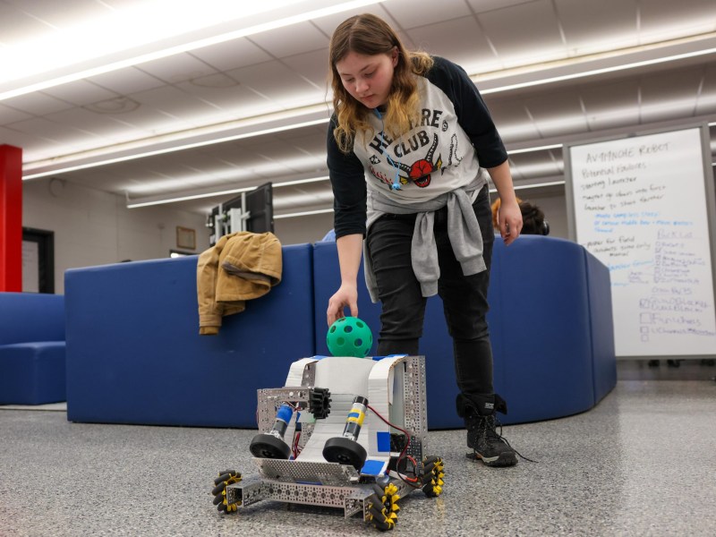 A person places a green perforated ball onto a small wheeled robot with metal framing inside a room with blue seating and a whiteboard.