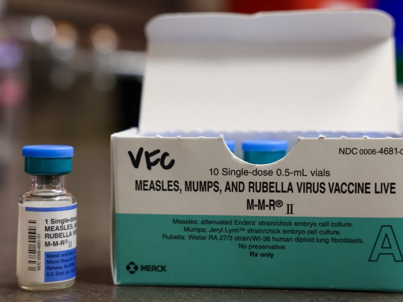 A vial and box labeled "Measles, Mumps, and Rubella Virus Vaccine Live M-M-R II" sit on a table, with "VFC" written on the box and blue-capped vials visible inside.