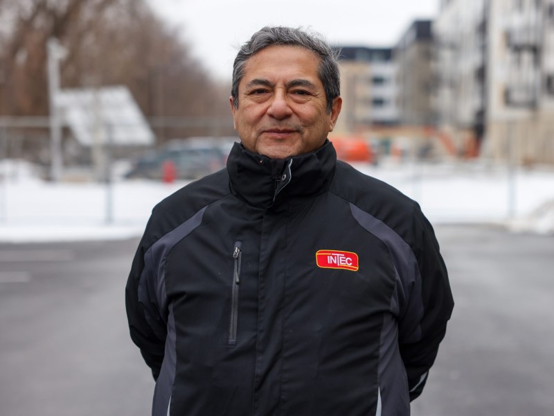 A person stands outdoors in a paved lot wearing a jacket with an "INTEC" logo, with snow, fencing and buildings out of focus in the background.