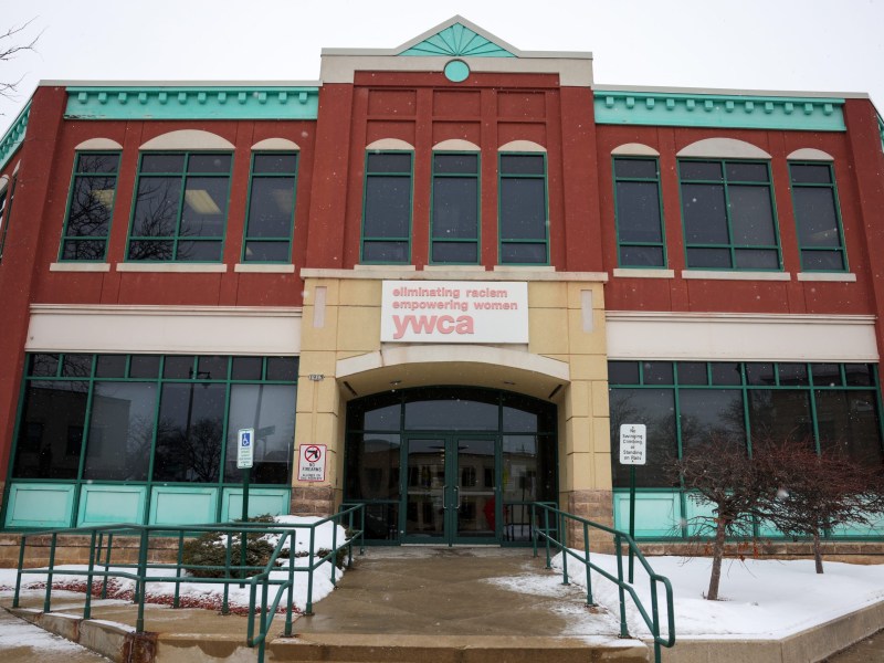A two-story brick building with green trim displays a sign reading “eliminating racism empowering women YWCA” above the entrance, with light snow on the ground in front.
