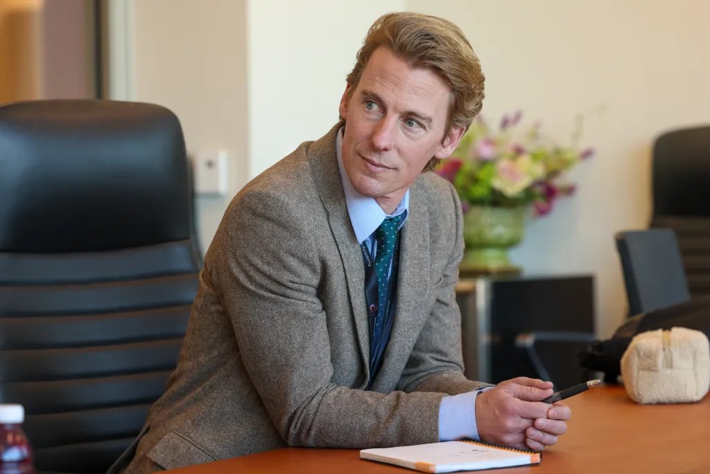 A person in a suit jacket and tie sits at a table and holds a pen next to a notebook, with a flower arrangement and chairs in the background.